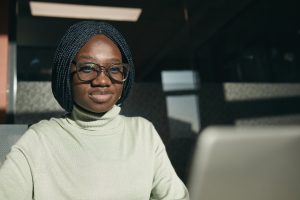 Confident woman with glasses and braided hair working at her laptop in a modern office setting.